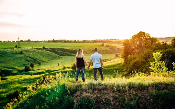 Girl and guy holding hands and standing on a green hill. Sunset. Back view. — Stock Photo, Image