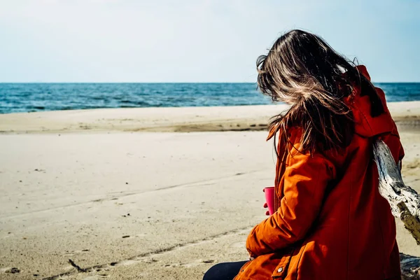 Junge schöne kaukasische Mädchen in einer roten warmen Jacke mit Kapuze, Schal, Lederhose sitzt auf einem felsigen Strand nach einem Spaziergang am Meer ausruhen — Stockfoto