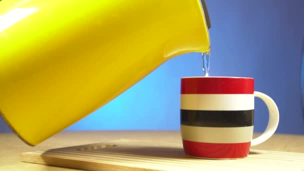 Chaleira elétrica amarela água fervente em uma xícara. A água fervida quente é derramada em uma caneca de copo de cerâmica vermelha, branca e azul  . — Vídeo de Stock