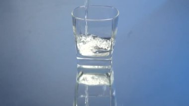 Şişeden kadehe votka dökme hareketi. Mavi arka planda bardağa votka dökülüyor. Ayna yansıması