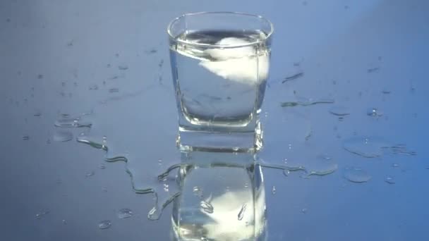 在玻璃杯中的伏特加中加入冰块 在蓝色背景中加入冰块 酒精透明冷冻饮料伏特加龙舌兰酒 慢动作镜像反射 — 图库视频影像