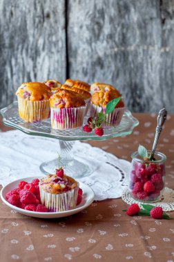 Böğürtlenlerle süslenmiş gri arka plandaki cam pasta standında ahududu kekleri, beyaz bir açık masa peçetesi ve kahverengi çiçekli masa örtüsü.