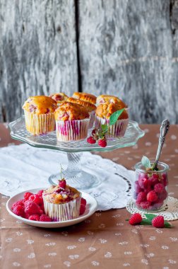 Böğürtlenlerle süslenmiş gri arka plandaki cam pasta standında ahududu kekleri, beyaz bir açık masa peçetesi ve kahverengi çiçekli masa örtüsü.