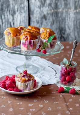 Böğürtlenlerle süslenmiş gri arka plandaki cam pasta standında ahududu kekleri, beyaz bir açık masa peçetesi ve kahverengi çiçekli masa örtüsü.