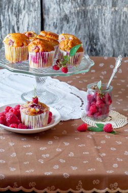 Böğürtlenlerle süslenmiş gri arka plandaki cam pasta standında ahududu kekleri, beyaz bir açık masa peçetesi ve kahverengi çiçekli masa örtüsü.
