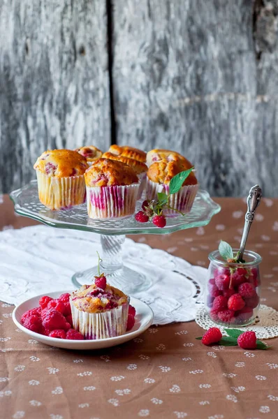 Böğürtlenlerle süslenmiş gri arka plandaki cam pasta standında ahududu kekleri, beyaz bir açık masa peçetesi ve kahverengi çiçekli masa örtüsü.