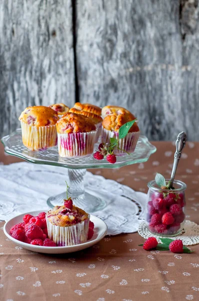 Böğürtlenlerle süslenmiş gri arka plandaki cam pasta standında ahududu kekleri, beyaz bir açık masa peçetesi ve kahverengi çiçekli masa örtüsü.