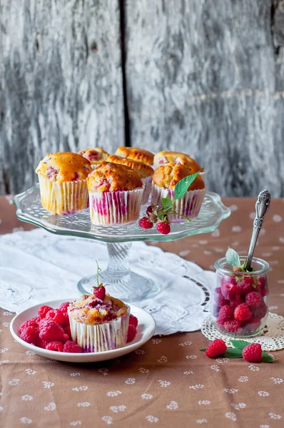 Böğürtlenlerle süslenmiş gri arka plandaki cam pasta standında ahududu kekleri, beyaz bir açık masa peçetesi ve kahverengi çiçekli masa örtüsü.