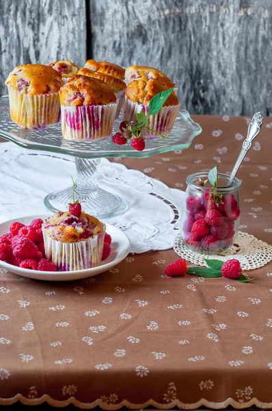 Böğürtlenlerle süslenmiş gri arka plandaki cam pasta standında ahududu kekleri, beyaz bir açık masa peçetesi ve kahverengi çiçekli masa örtüsü.