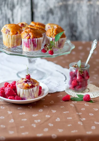 Böğürtlenlerle süslenmiş gri arka plandaki cam pasta standında ahududu kekleri, beyaz bir açık masa peçetesi ve kahverengi çiçekli masa örtüsü.