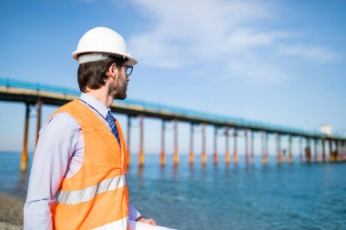 Şapkalı genç adam inşaatta çalışıyor. Deniz kenarında köprü tadilatında çalışan mimar.