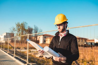 Şapkalı genç adam inşaatta çalışıyor. Kentsel arkaplan üzerine plan üzerinde çalışan mimar.