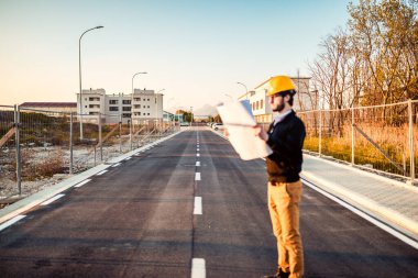 Şapkalı genç adam inşaatta çalışıyor. Kentsel arkaplan üzerine plan üzerinde çalışan mimar.