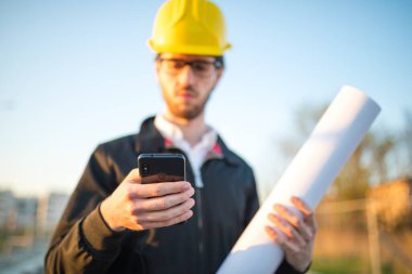 Şapkalı genç adam inşaatta çalışıyor. Kentsel arkaplan üzerine plan üzerinde çalışan mimar.