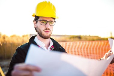 Şapkalı genç adam inşaatta çalışıyor. Kentsel arkaplan üzerine plan üzerinde çalışan mimar.