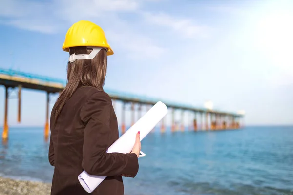 Young woman in hard hat working on construction. Architect working with bridge renovation by the seaside.