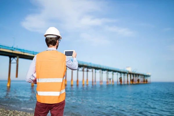 Young man in hard hat working on construction. Architect working with bridge renovation by the seaside.