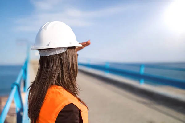 Young woman in hard hat working on construction. Architect working with bridge renovation by the seaside.
