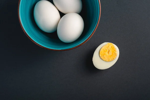 Boiled egg with yellow yolk near to white eggs in blue bowl on dark moody black plain minimal background, top view, happy Easter day