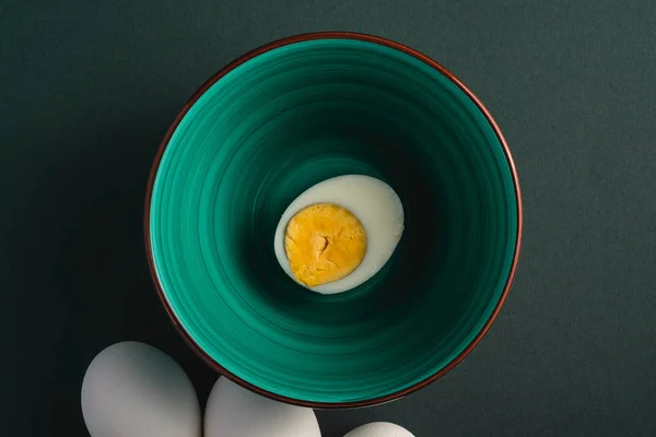 Boiled egg with yellow yolk in green bowl near to white eggs on dark moody black plain minimal background, top view, happy Easter day