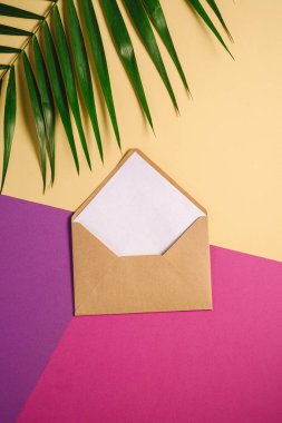 Palmiye yaprakları üzerinde beyaz boş kart, pembe, mor ve krem sarısı arkaplan, sahte boş mektup içeren Kraft kahverengi kağıt zarf