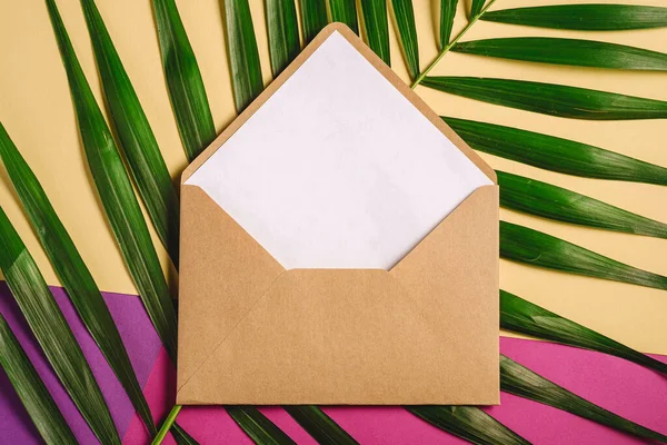 Kraft brown paper envelope with white empty card on palm leaves, pink, purple and cream yellow background, mockup blank letter