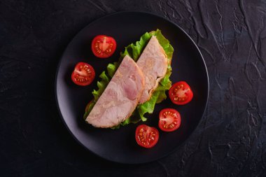 Hindi jambonlu sandviç, yeşil salata ve siyah tabakta taze vişneli domates dilimleri, koyu dokulu arka plan, üst manzara.