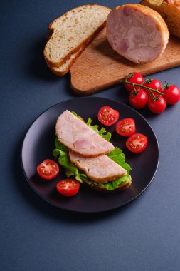 Hindi jambonlu sandviç, yeşil salata ve taze vişneli domates dilimleri kesme tahtasındaki malzemelerin yanında, mavi minimal arka plan, açı görüşü.