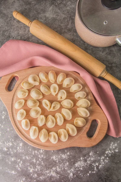 Raw dumplings on a wooden round board. Near a rolling pin, saucepan, scattered flour - top view