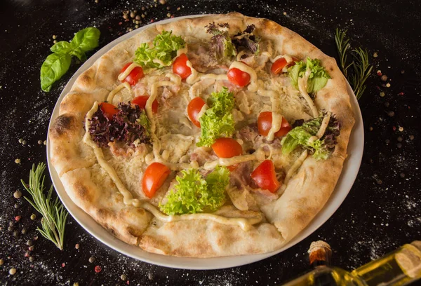 Sezar soslu pizza, et, rendelenmiş peynir, doğranmış domates ve salata. Baharat ve malzemelerle serpiştirilmiş siyah bir masanın üzerinde. Menü için fotoğraf.