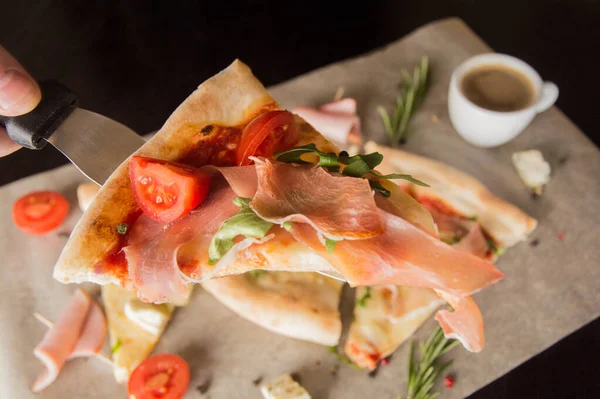 A slice of pizza with prosciutto - on a pizza spatula. In the background pizza and coffee.
