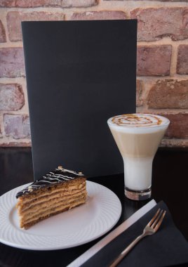Beyaz tabakta bir parça kek - uzun bir bardakta latte ile siyah bir masada menü, kırmızı tuğlalı bir duvarın arka planında.