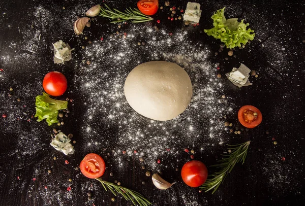 Pizza hamuru, beyaz un ve pizza malzemeleriyle siyah bir masanın üzerinde daire şeklinde dizilmiş. Yarı bitmiş pizza hamuru..