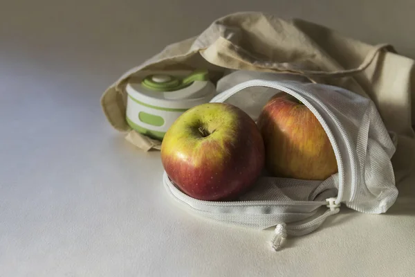 Shopper bag with collapsible silicone mug and apples in reusable fruit pouch on white background in the sunlight with shadows. Concept of trendy environmental-friendly reusable stuff. Copy space