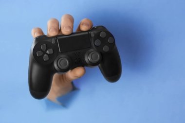 Erkeklerin eli video oyunu kumandasını yırtık mavi kağıt duvardaki delikten tutuyor. Kopya alanı ve seçici odaklı yatay resim. Evde kal konsepti 