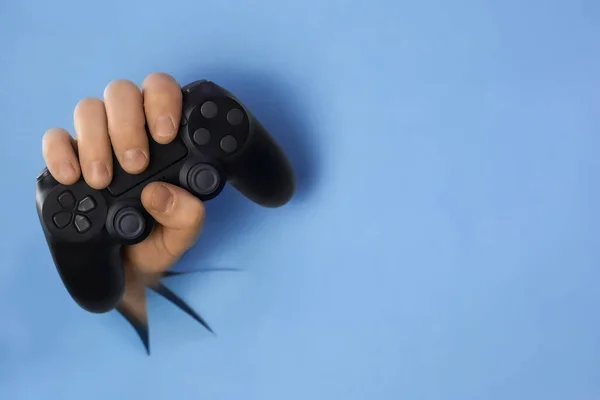男の手は青い紙の壁に穴を通ってビデオゲームコントローラーを保持しています コピースペースと選択フォーカス付きの水平画像 家のコンセプトを守る — ストック写真