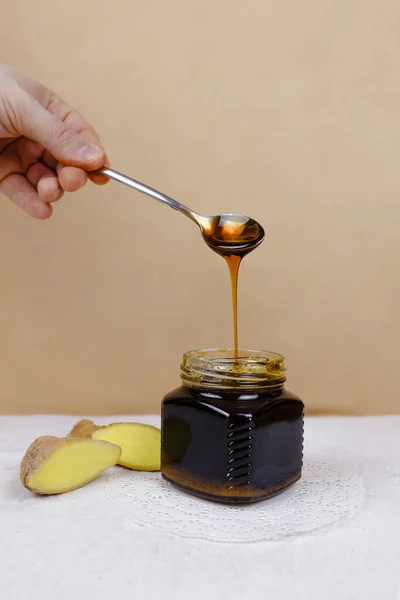 Buckwheat honey pours from spoon into jar and sliced ginger lying near. Natural medicine, healthy food, ethnoscience, strengthening immunity. Vertical picture, close-up, selective focus.