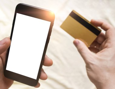 İnternetten alışveriş konsepti. Abonelikleri öde. Akıllı telefon ve kredi kartı adamın elinde. Arka planda buruşuk yatak. Evde kalıp karantinaya alın. Çevrimiçi çağrı. Teletıp. Beyaz boş ekran maketi