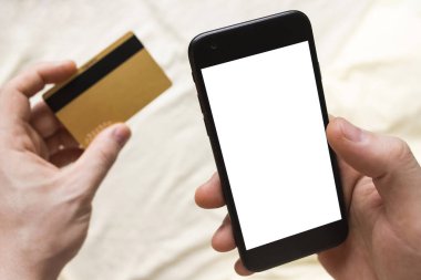 İnternetten alışveriş konsepti. Abonelikleri öde. Akıllı telefon ve kredi kartı adamın elinde. Arka planda buruşuk yatak. Evde kalıp karantinaya alın. Çevrimiçi çağrı. Teletıp. Beyaz boş ekran maketi