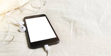 Kulaklıklı akıllı telefon buruşuk yatakta yatıyordu. Yakın plan, sığ alan derinliği. Karantinaya alınırken evde kal. Mesafe öğrenme. Çevrimiçi çağrı. Uzaktan kumanda. Beyaz maket boş ekran. 