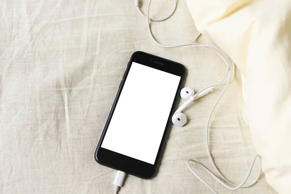 Kulaklıklı akıllı telefon buruşuk yatakta yatıyordu. Karantinaya alınırken evde kal. Mesafe öğrenme. Çevrimiçi çağrı. Uzaktan kumanda. Beyaz maket boş ekran.