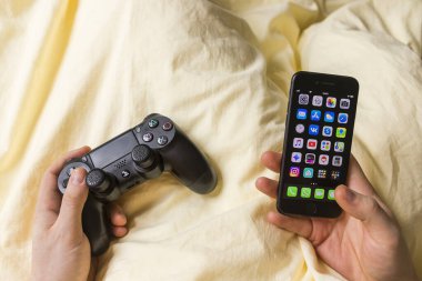SAINT-PETERSBURG RUSSIA 19 Nisan 2020: Dualshock 4 denetleyici ve Apple iPhone ve ekrandaki uygulamalar erkeklerin elinde. Oyuncu adam buruşuk yatakta oturur. Kumar bağımlılığı konsepti. Evde kal..