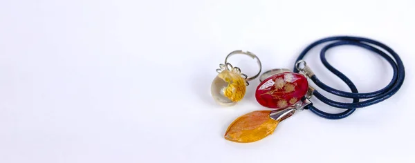 Beyaz zemin üzerinde epoksi reçineden yapılmış kristallerin içinde kurumuş çiçekler olan kolye ve yüzükler. Yakın çekim alan derinliği. El yapımı, DIY bijouterie düz pankart