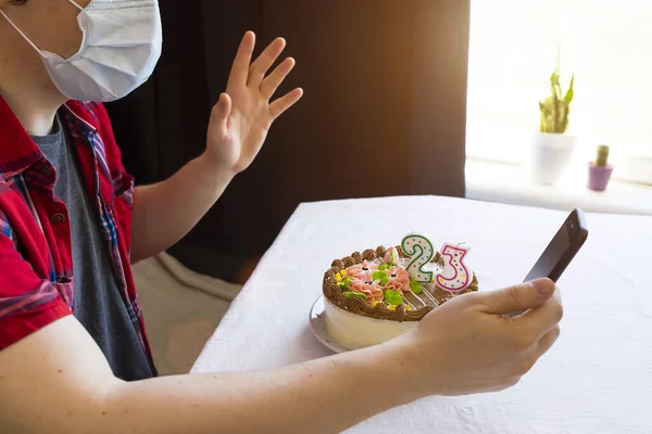 Lone quarantine birthday party. Man in medical mask sits with a smartphone in his hands. Birthday cake with candles. Stay home while quarantine. Online call. Video conference with friends or parents.