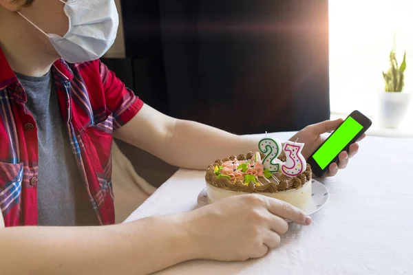 Quarantine birthday. Man in medical mask sits with smartphone in his hand. Birthday cake with candles. Online call. Video conference with friends or parents. Green mock-up blank screen.