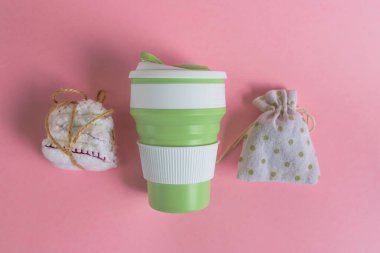 Katlanabilir silikon bardak, ağ torbası ve sicimle tutturulmuş yüz veya makyaj malzemesi için yeniden kullanılabilir pamuk diskler. Pembe arka planda sıfır atık kavramı üst görünümü. Çevre dostu yaşam tarzı 