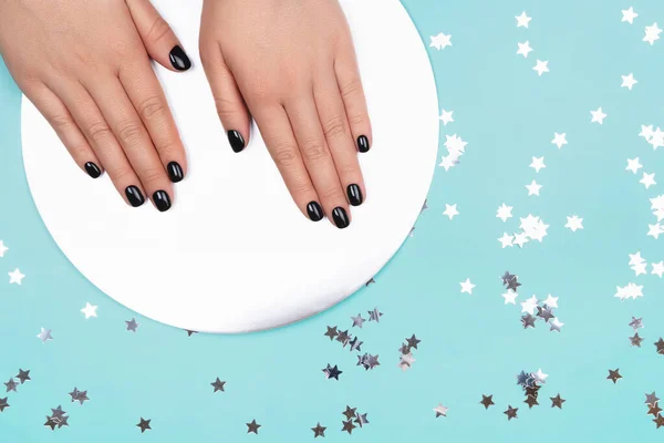 Womans hands with black manicure on silver circle and pastel blue background.