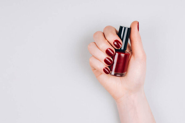 Beautiful dark red manicure with a bottle of nail polish in hand on a grey background. Flat lay style.