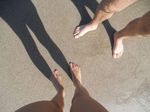 Vue de la section basse d'un homme et des femmes jeunes jambes de couple debout sur le sable sur la plage à midi. Ombres des jambes sur le sable. — Photo