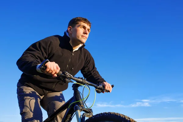 Retrato de ciclista de montanha, ciclista de jaqueta preta com bicicleta de montanha, céu azul no fundo, vista inferior . — Fotografia de Stock